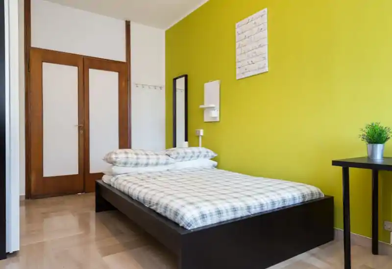 Bright, modern bedroom with a double bed on a low dark frame against a vibrant green accent wall; plaid bedding, bedside mirror and small desk with plant complete the minimalist, welcoming setting.