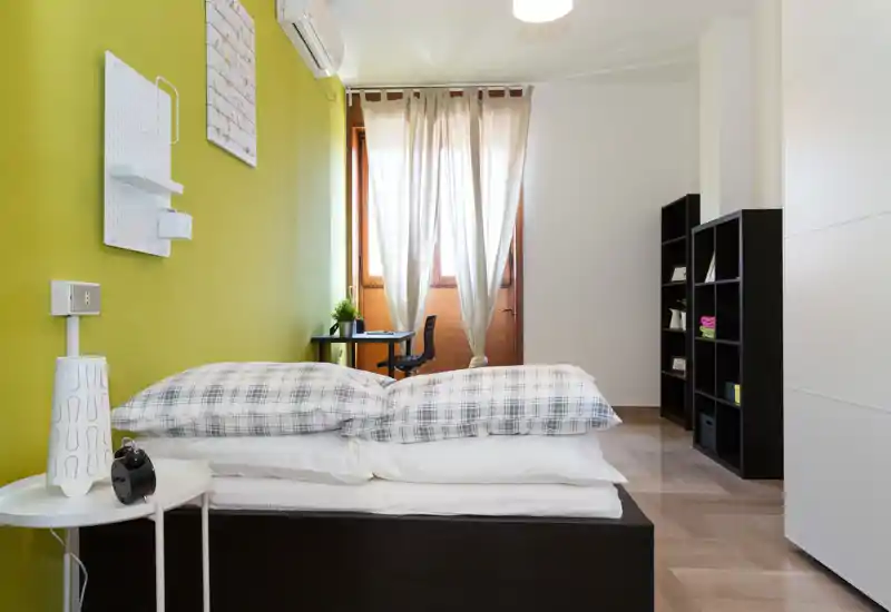 Bright, modern bedroom with a neatly made double bed in the foreground, green accent wall, desk by the window and black shelving for storage.