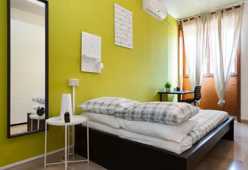 Dormitorio moderno y luminoso con cama baja, ropa de cama a cuadros y un rincón de trabajo junto a la ventana soleada. La pared verde y la decoración minimalista ofrecen una atmósfera fresca y acogedora.