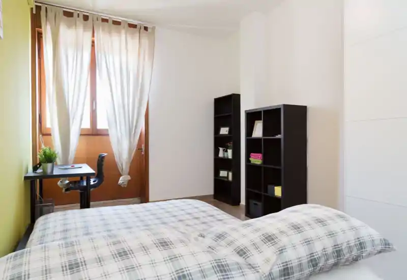 Dormitorio luminoso y ordenado con cama doble en primer plano, escritorio junto a una gran ventana con cortinas translúcidas y estanterías abiertas para almacenamiento. La luz natural y los tonos neutros hacen la habitación agradable y relajante.