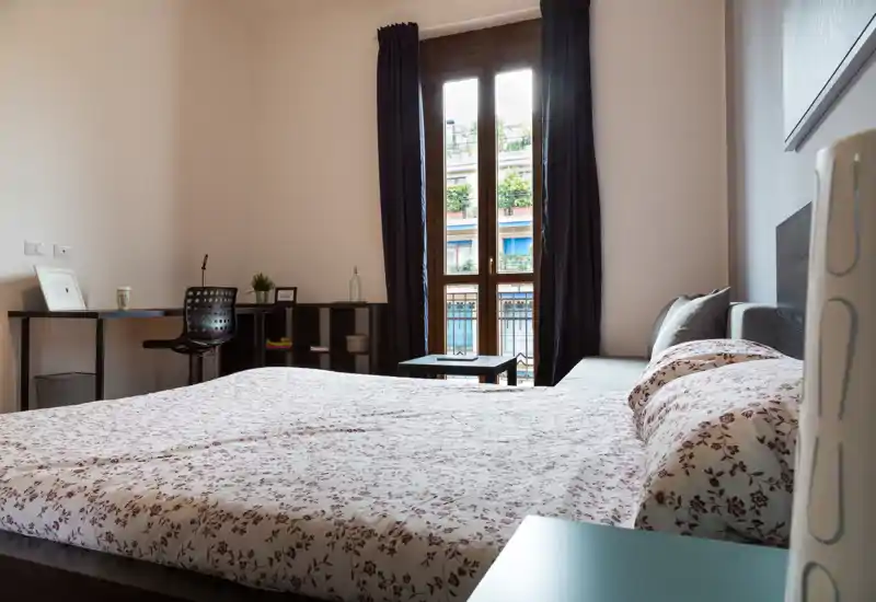 Dormitorio luminoso y bien decorado con una cama grande en primer plano, ropa de cama estampada y una ventana alta que aporta mucha luz natural.