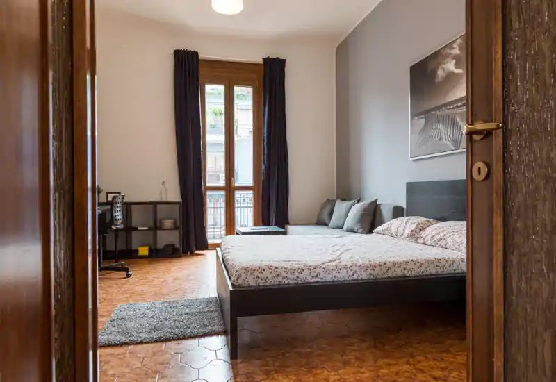 Dormitorio luminoso y bien decorado con cama doble, cojines en el sofá y luz natural desde la puerta del balcón — tonos neutros y suelo de madera para un ambiente acogedor urbano.