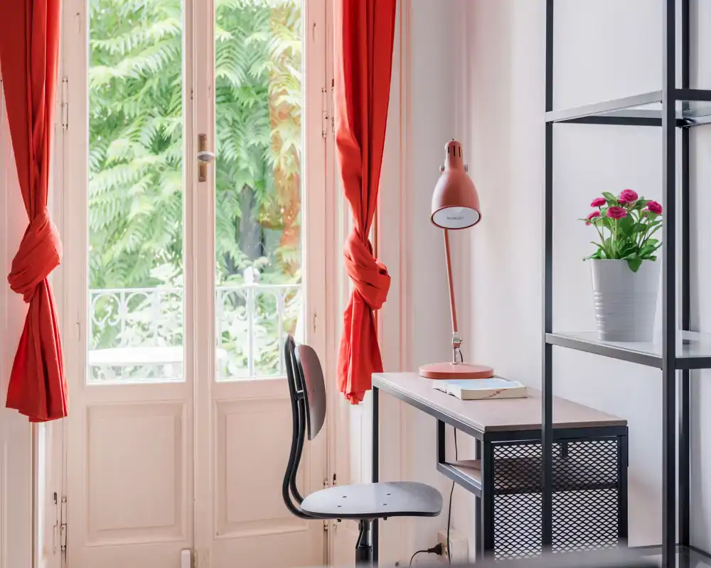 Pequeño espacio de trabajo luminoso junto a las puertas-fenétrales con cortinas rojas, escritorio estrecho, lámpara de estudio y planta en maceta — ideal para teletrabajo con abundante luz natural.