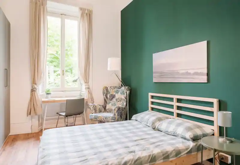 Dormitorio luminoso y bien decorado con cama de lamas de madera, ropa de cama a cuadros y pared verde; incluye un acogedor sillón estampado y un pequeño escritorio junto a la ventana.