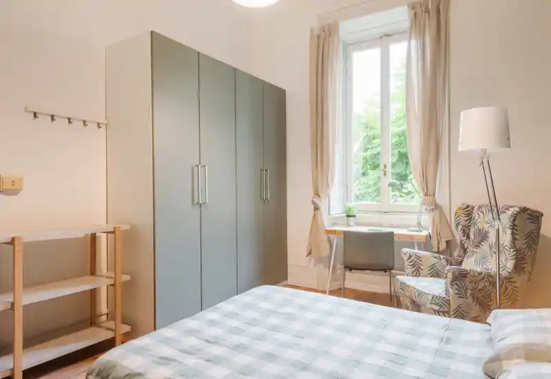 Dormitorio luminoso y bien presentado con cama doble en primer plano, amplio armario, escritorio junto a una ventana alta y un sillón estampado — perfecto para una estancia cómoda con mucha luz natural.