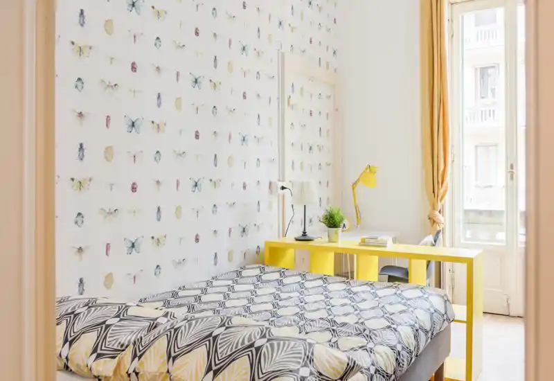 Dormitorio luminoso y con estilo, papel pintado de mariposas, edredón estampado y un escritorio amarillo junto a una gran ventana que aporta mucha luz natural.