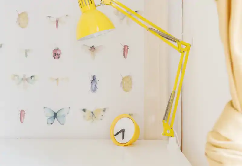 Primer plano de una superficie de escritorio o mesita clara con una lámpara articulada amarilla y un reloj despertador amarillo a juego; pegatinas de insectos y mariposas en la pared aportan un toque decorativo y divertido.