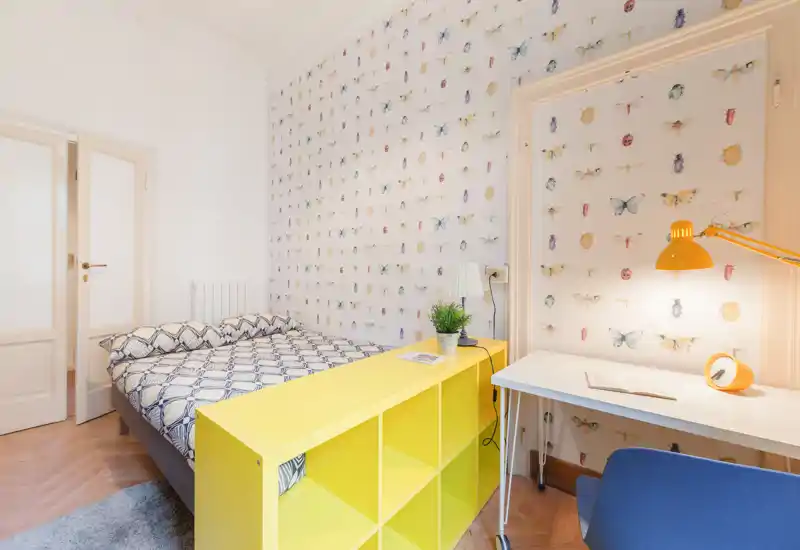 Dormitorio luminoso y bien decorado con cama completa, papel pintado con estampado y una pequeña zona de estudio — ideal para viajeros solos o parejas que buscan un espacio acogedor y colorido.