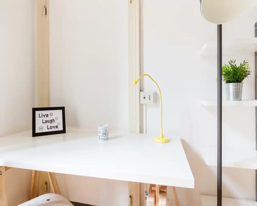 Espace de travail minimal et lumineux avec bureau blanc en coin, petite lampe jaune, cadre décoratif et plante en pot — idéal pour le télétravail ou les études.