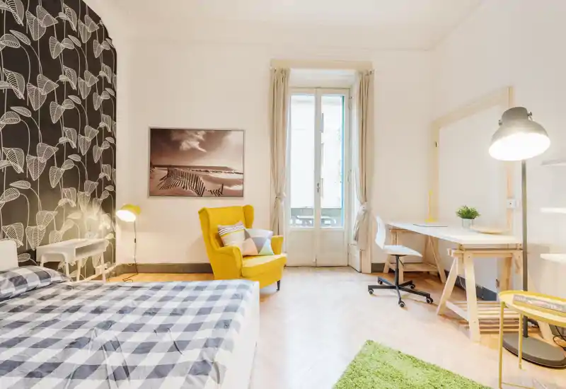 Chambre lumineuse et moderne avec un grand lit à carreaux, un fauteuil jaune et un espace bureau près de la fenêtre — décoration chaleureuse et idéale pour une annonce locative.