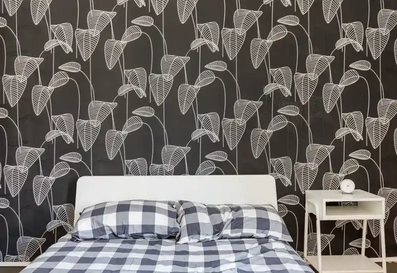Chambre lumineuse et décorée avec un grand lit aux couettes à carreaux, un papier peint graphique derrière la tête de lit et une petite table de chevet blanche avec réveil.