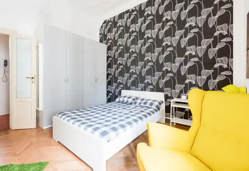 Chambre lumineuse et moderne avec lit double blanc, papier peint sombre à motifs et un fauteuil jaune confortable — idéale pour mettre en avant la décoration contemporaine et la lumière naturelle.