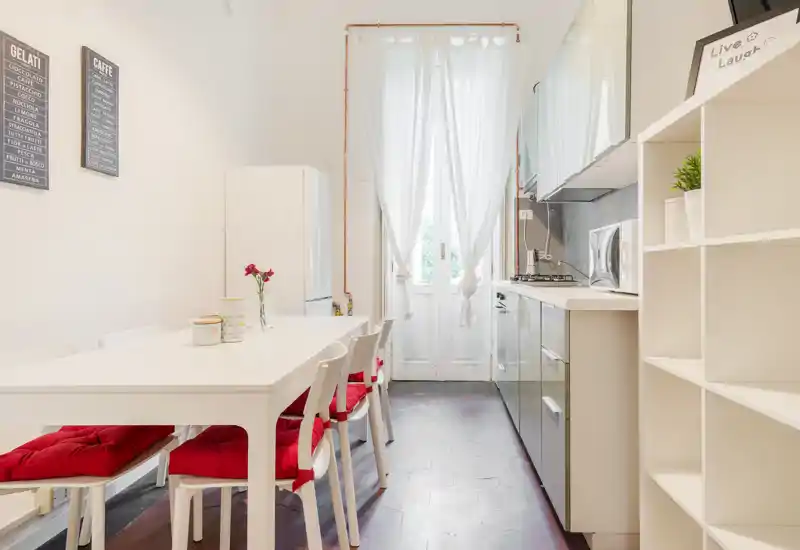Cocina moderna y luminosa con una amplia mesa blanca y cojines rojos; la luz natural entra por la puerta acristalada y los muebles y electrodomésticos ocupan una pared.