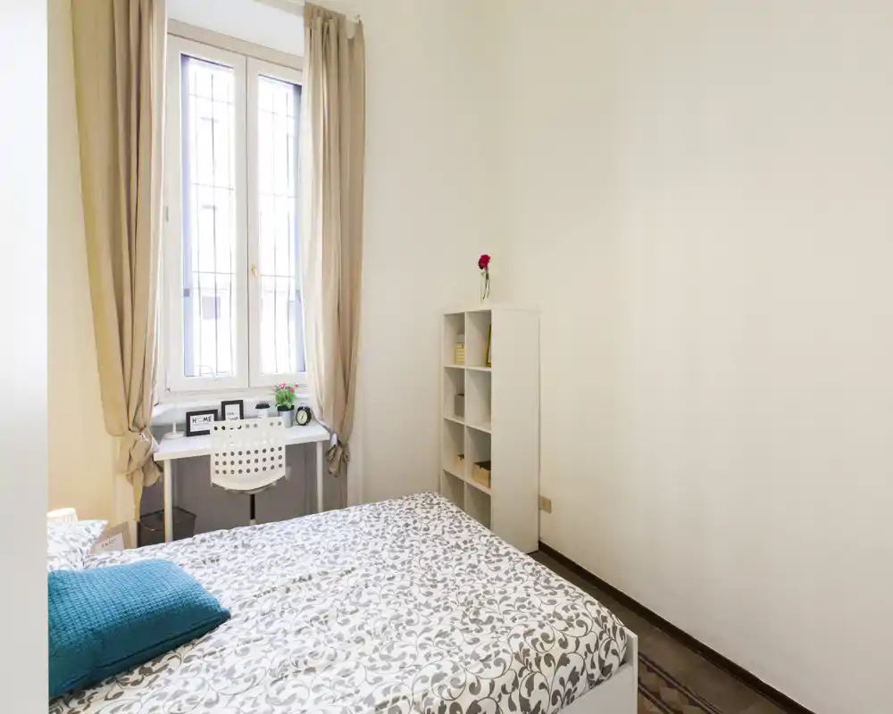Dormitorio luminoso y ordenado con cama doble con estampado, un pequeño escritorio junto a la ventana y una estantería estrecha — ideal para un alquiler compacto y bien iluminado.