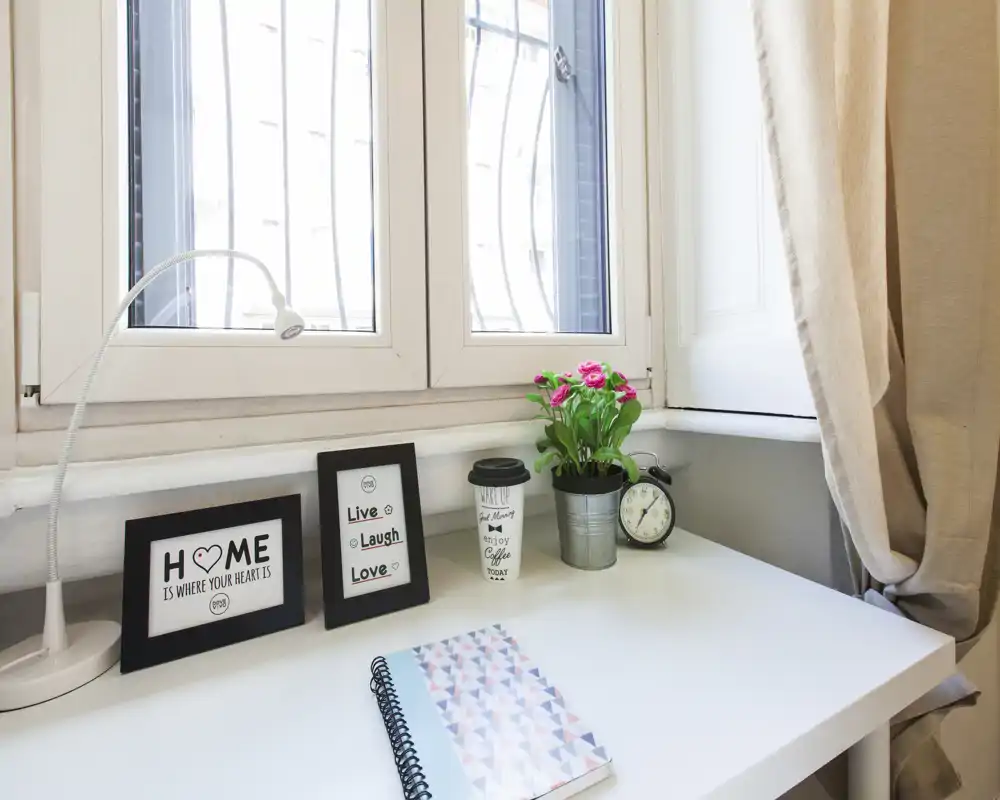 Rincón de escritorio luminoso junto a la ventana con marcos decorativos, una pequeña planta y un cuaderno: un espacio de trabajo compacto y ordenado con luz natural.