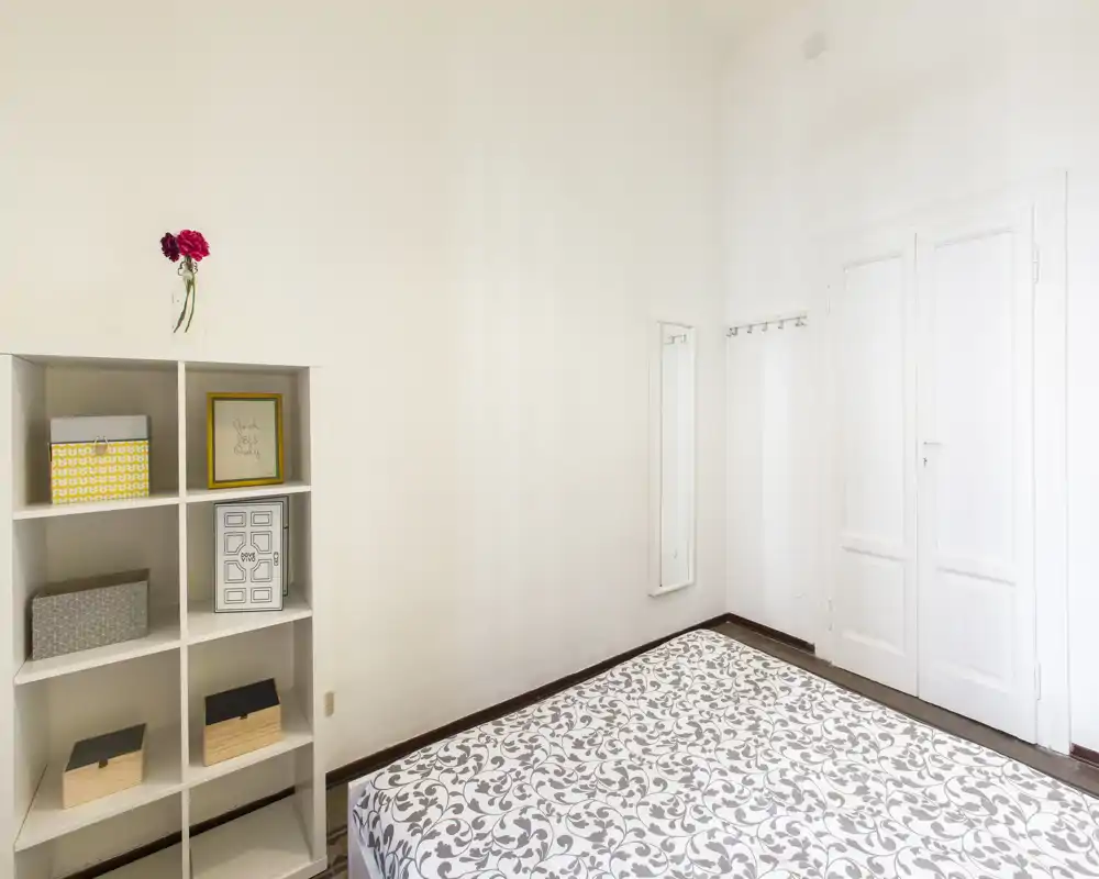 Dormitorio luminoso y minimalista con colchón estampado visible, una estantería con cajas y paredes y puertas blancas que crean una sensación limpia y aireada.
