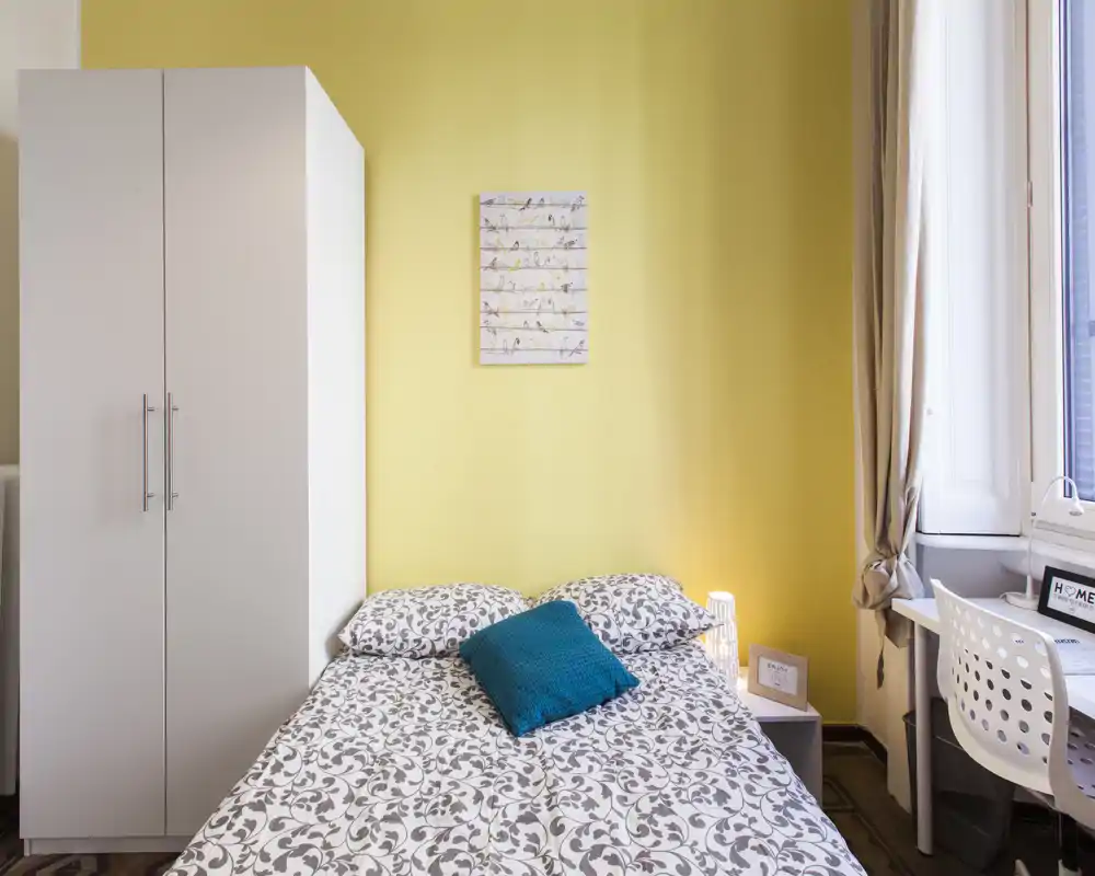 Dormitorio luminoso y ordenado con cama doble, ropa de cama estampada y cojín turquesa. Un armario blanco y un escritorio junto a la ventana completan la distribución, perfecto para un anuncio de alquiler.