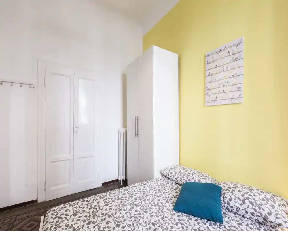 Dormitorio luminoso y acogedor con ropa de cama estampada, cojín turquesa y armario blanco sobre pared amarilla — perfecto para un anuncio de alquiler.