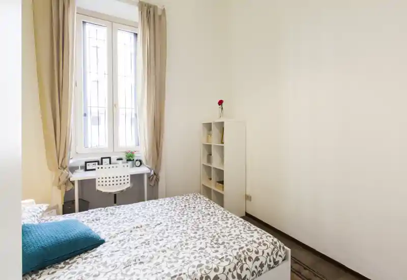 Dormitorio luminoso y ordenado con cama doble con estampado, un pequeño escritorio junto a la ventana y una estantería estrecha — ideal para un alquiler compacto y bien iluminado.