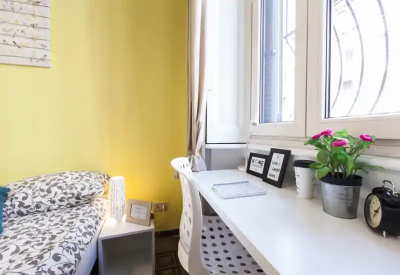 Rincón de dormitorio luminoso y bien decorado con cama estampada junto a un escritorio blanco bajo una gran ventana; lámpara, marcos decorativos y planta pequeña crean un ambiente acogedor y brillante.