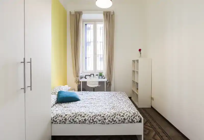 Dormitorio luminoso y bien decorado con cama doble, ropa de cama estampada y un pequeño escritorio junto a la ventana; paredes neutras y un toque de color que crean un ambiente acogedor.