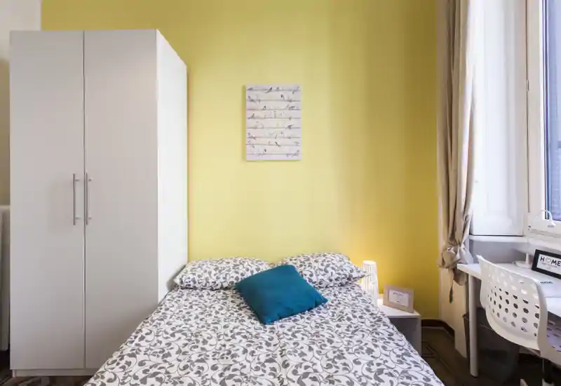 Dormitorio luminoso y ordenado con cama doble, ropa de cama estampada y cojín turquesa. Un armario blanco y un escritorio junto a la ventana completan la distribución, perfecto para un anuncio de alquiler.