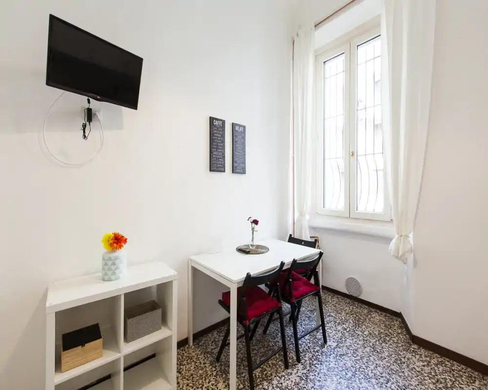 Pequeño rincón comedor luminoso con mesa blanca, dos sillas plegables negras con cojines rojos y una ventana que aporta luz natural. Toques decorativos mínimos que crean un espacio ordenado y acogedor, ideal para comidas informales.