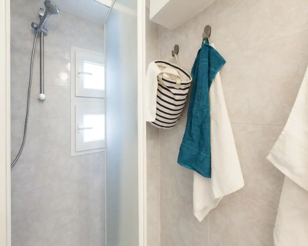 Ducha de un baño pequeño y luminoso con teleducha montada en la pared, mampara de vidrio esmerilado y toallas colgadas en ganchos.