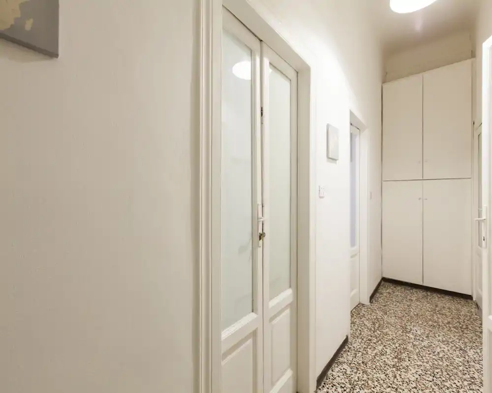 Entrada luminosa y pasillo estrecho con puertas blancas y armarios empotrados, suelo de terrazo e iluminación suave en el techo.