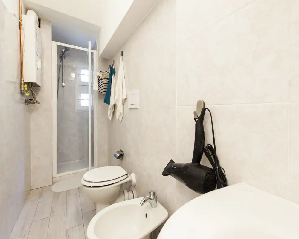 Baño compacto y luminoso con cabina de ducha, inodoro, bidé y secador de pelo montado. Azulejos neutros y suelo efecto madera para un espacio limpio y funcional.