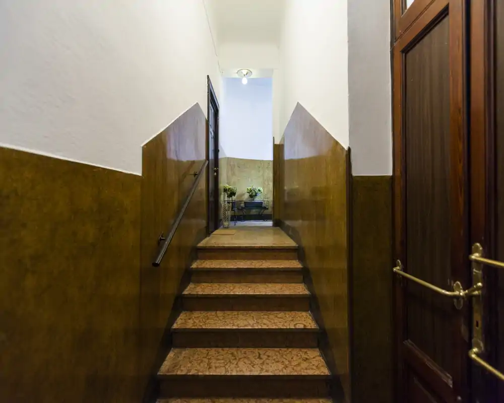 Una estrecha escalera de entrada bien iluminada con zócalos de madera en las paredes inferiores, pasamanos metálico y un pequeño rellano que conduce a un pasillo interior. Los tonos cálidos y la iluminación equilibrada crean una primera impresión acogedora.