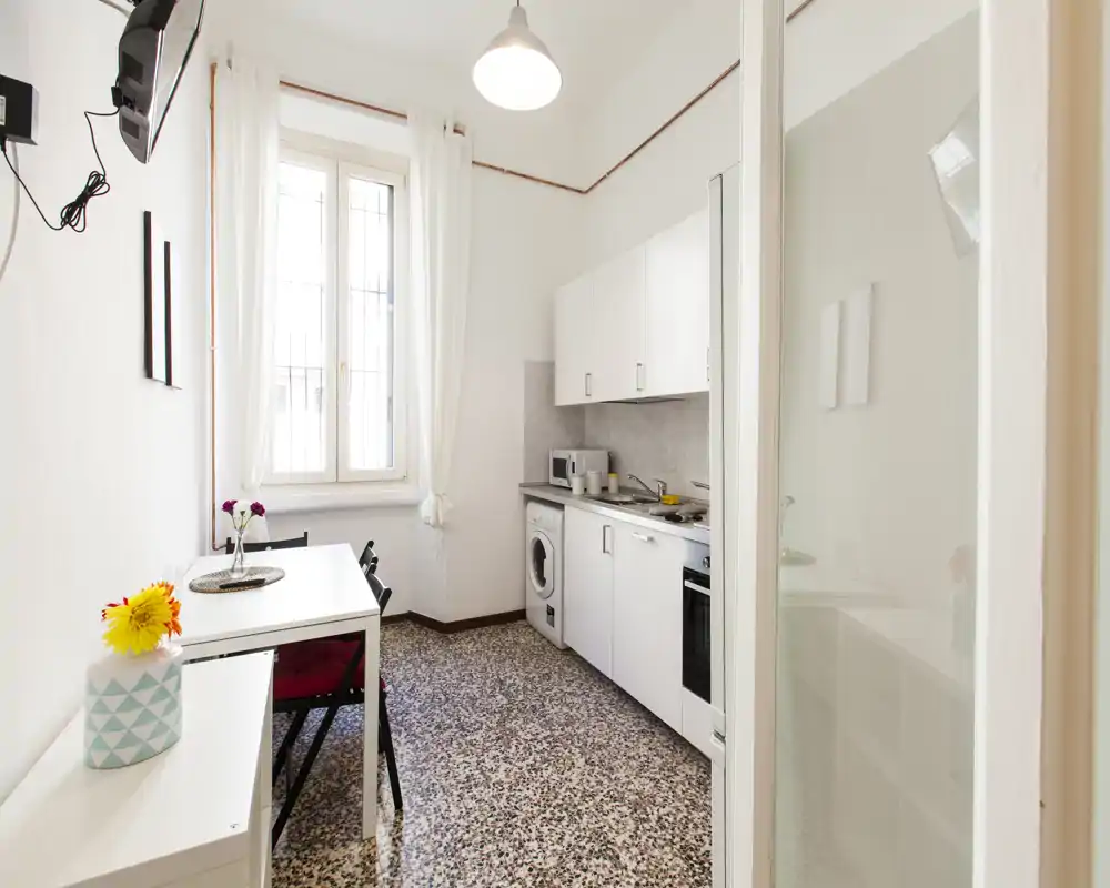 Cocina compacta y luminosa con armarios blancos, pequeña mesa de comedor y luz natural — incluye lavadora y horno, ideal para un apartamento urbano acogedor.