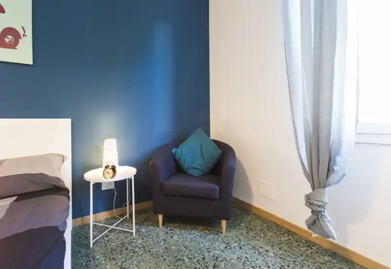 Rincón de dormitorio acogedor con borde de la cama visible, una mesita redonda con lámpara y despertador, un sillón cómodo y luz natural desde la ventana.