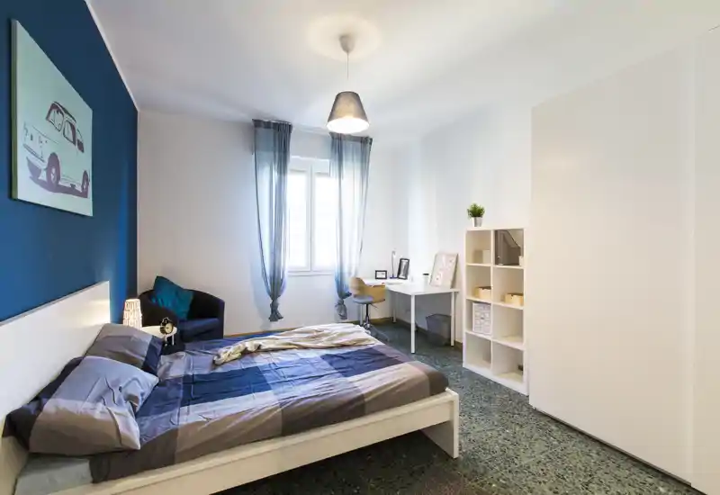 Bright, modern bedroom with a double bed framed by a deep blue accent wall, soft curtains and a small workspace. The room feels tidy and welcoming with contemporary furnishings and good natural light.