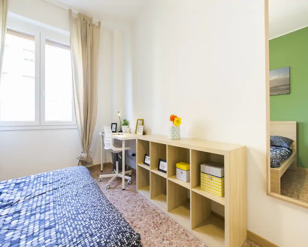 Camera da letto luminosa e ben allestita con copriletto a fantasia, scrivania vicino alla finestra e scaffalatura modulare per contenere oggetti — ideale per uno studente o una persona sola.