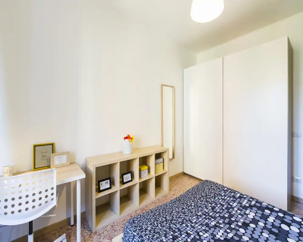 Camera da letto luminosa e minimal con copriletto a fantasia, armadio bianco e libreria in legno chiaro; scrivania e sedia offrono un comodo angolo lavoro.