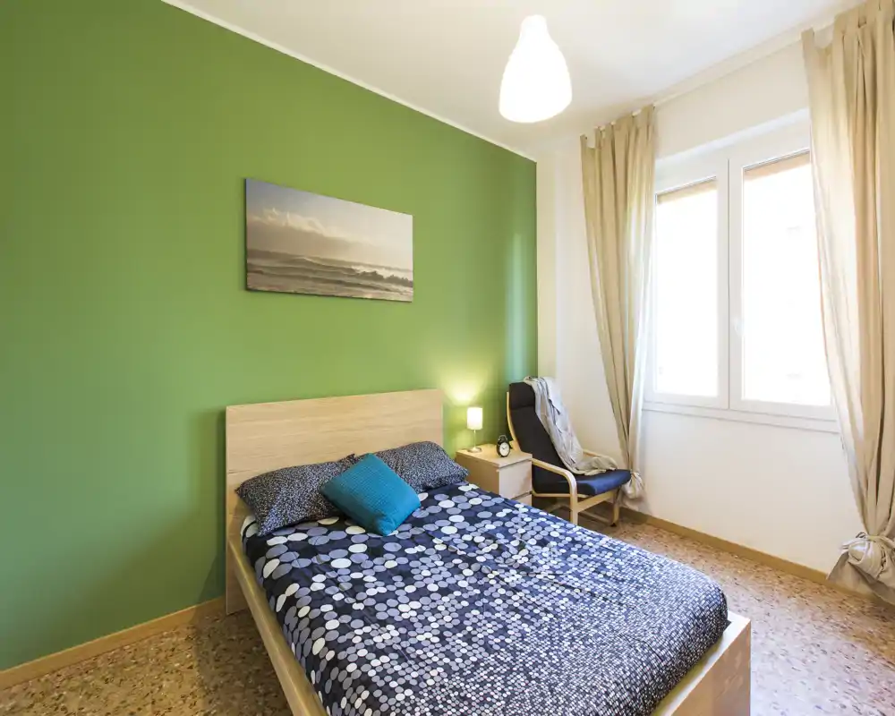 Camera da letto luminosa e ordinata con testiera in legno, biancheria a fantasia e parete verde; molta luce naturale da una grande finestra e una poltrona per la lettura accanto al comodino.