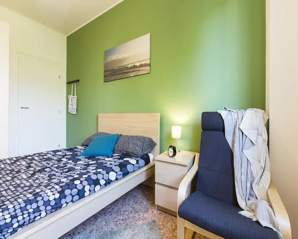 Camera da letto luminosa e moderna con struttura letto in legno chiaro, biancheria a fantasia e una poltrona accogliente vicino a una parete verde. Comodino con lampada e decorazioni semplici e curate.