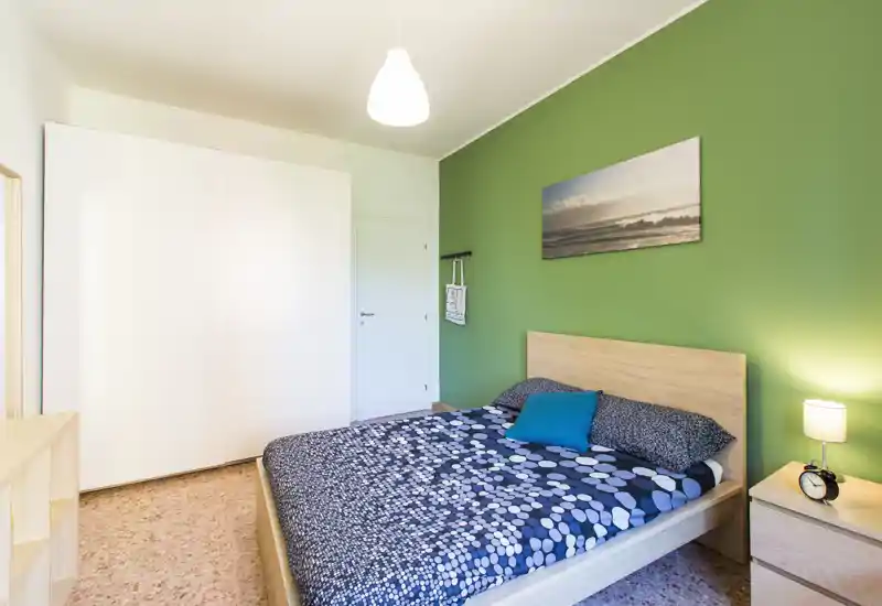 Camera da letto moderna e luminosa con letto matrimoniale, comodino e armadio; parete verde e biancheria blu coordinata creano un’atmosfera accogliente e fresca, perfetta per annunci.
