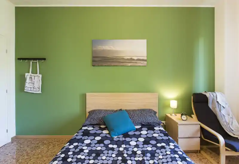 Camera da letto luminosa e ben curata con letto matrimoniale su una parete verde, comodino con lampada e una poltrona accogliente vicino alla finestra.