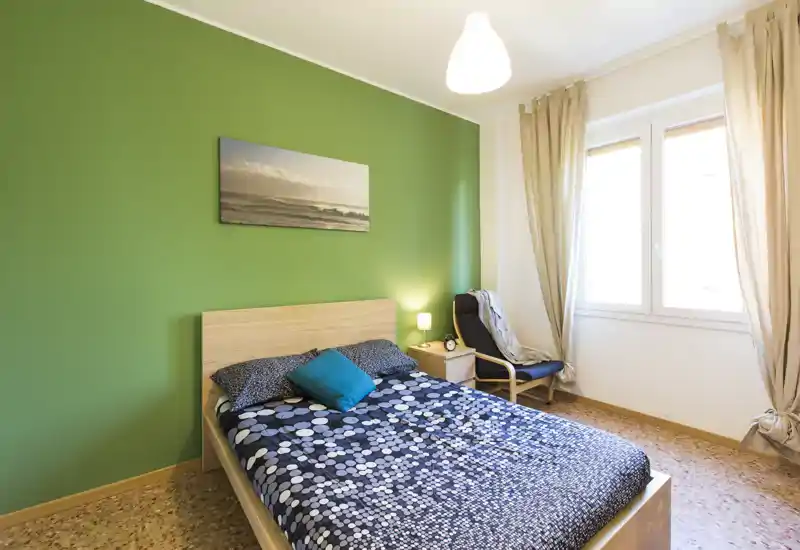 Bright, neatly styled bedroom with a wood headboard, patterned bedding and a green accent wall; natural light from a large window and a cozy reading chair by the bedside.