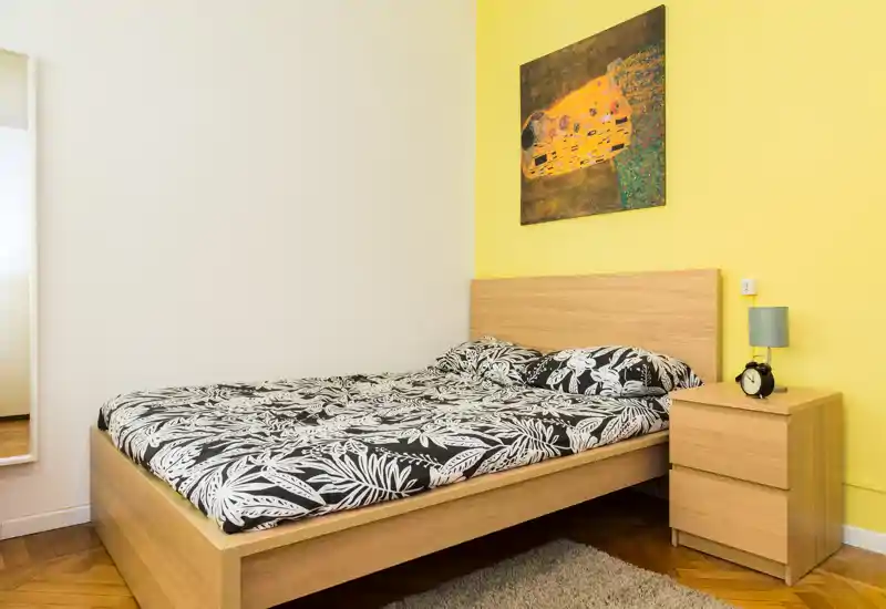 Camera da letto luminosa e ben arredata con letto in legno, biancheria a fantasia, comodino con lampada; parete gialla e quadro donano carattere.