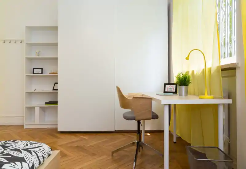 Angolo camera luminoso e moderno con letto in ordine, armadio bianco e una scrivania minimalista vicino a una finestra soleggiata, ideale per lavorare e riposare.