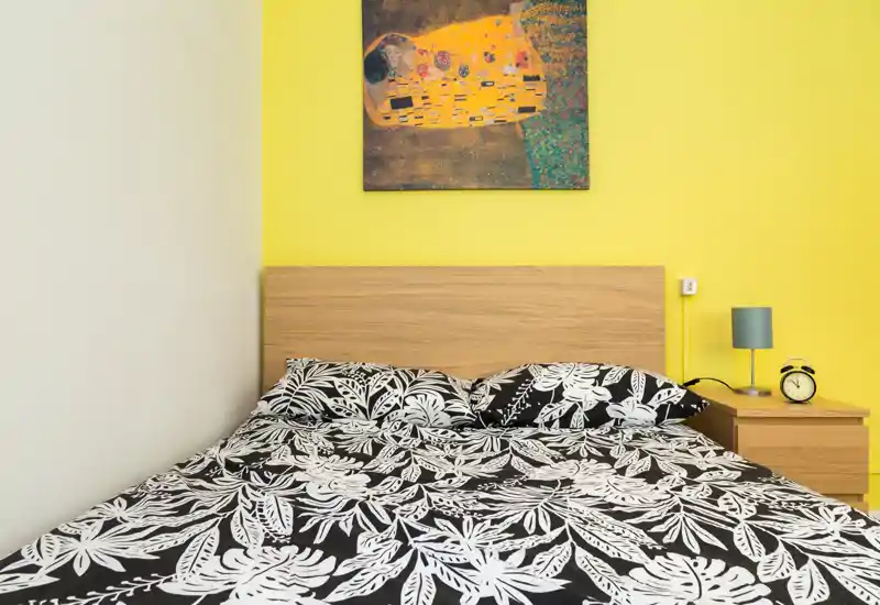 Camera da letto moderna e luminosa con testiera in legno, biancheria nera e bianca a motivo, comodino con lampada e sveglia e un dipinto colorato sopra il letto.