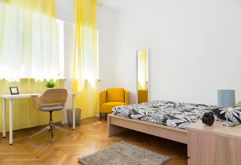 Camera da letto luminosa e moderna con letto su pedana in legno, biancheria a motivo, poltrona gialla come elemento d'accento e ampie finestre con tende gialle leggere che filtrano molta luce naturale.