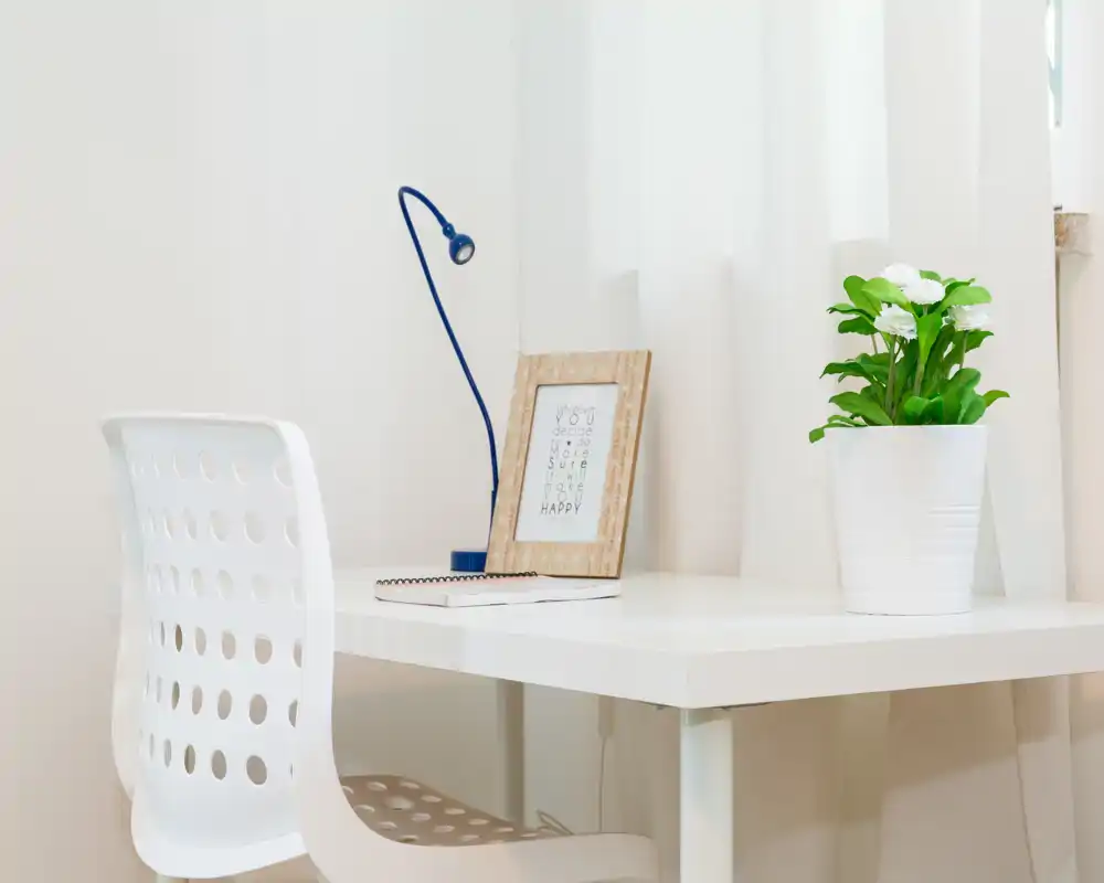 Angolo studio luminoso e minimale con scrivania bianca e sedia traforata, arricchito da una lampada da tavolo blu, una cornice e una pianta in vaso.