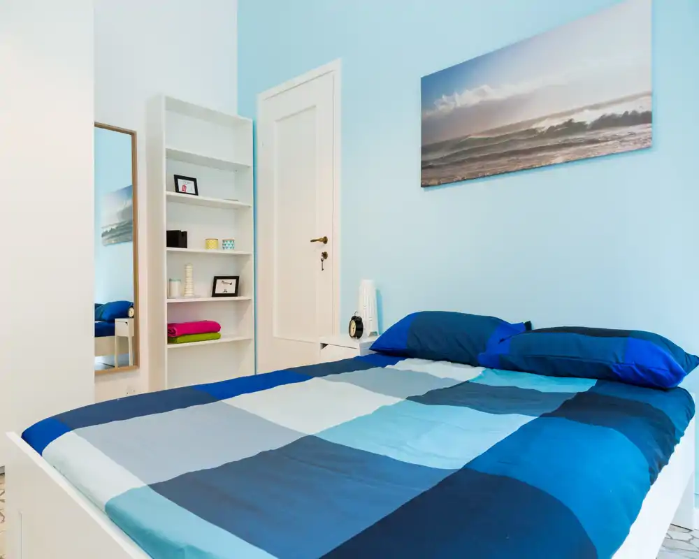 Camera da letto luminosa e ordinata con un ampio letto matrimoniale con lenzuola blu a quadrati, comodino semplice e quadro con tema marino.