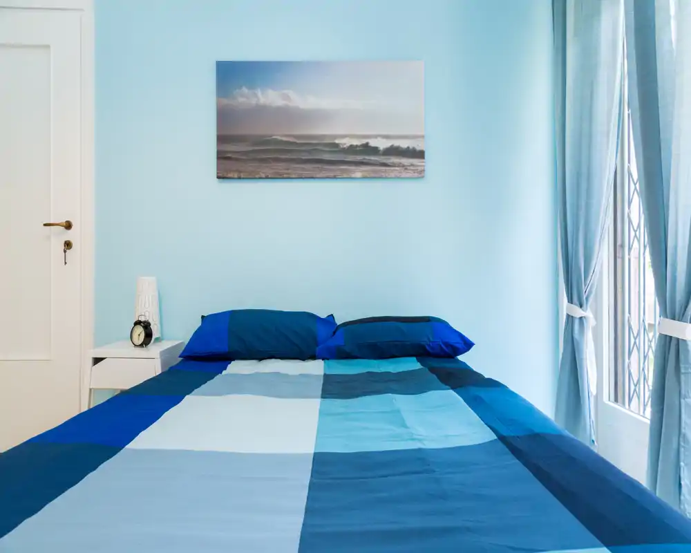 Camera da letto luminosa e ordinata con letto matrimoniale in tonalità di blu, comodino e luce naturale dalla finestra; un quadro con tema marino dona un tocco rilassante.