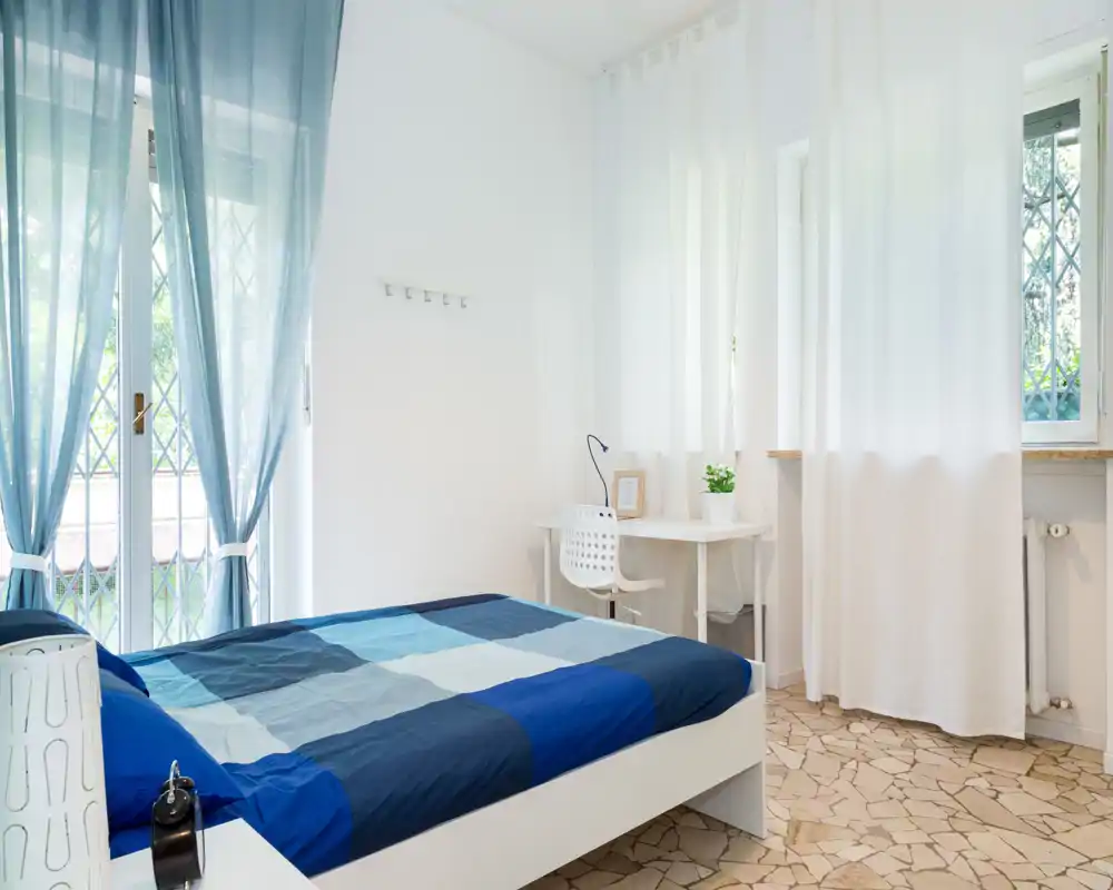 Camera da letto luminosa e minimal con matrimoniale in toni blu, ampie finestre con tende leggere e una scrivania vicino alla finestra — ideale per soggiorni confortevoli e tanta luce naturale.