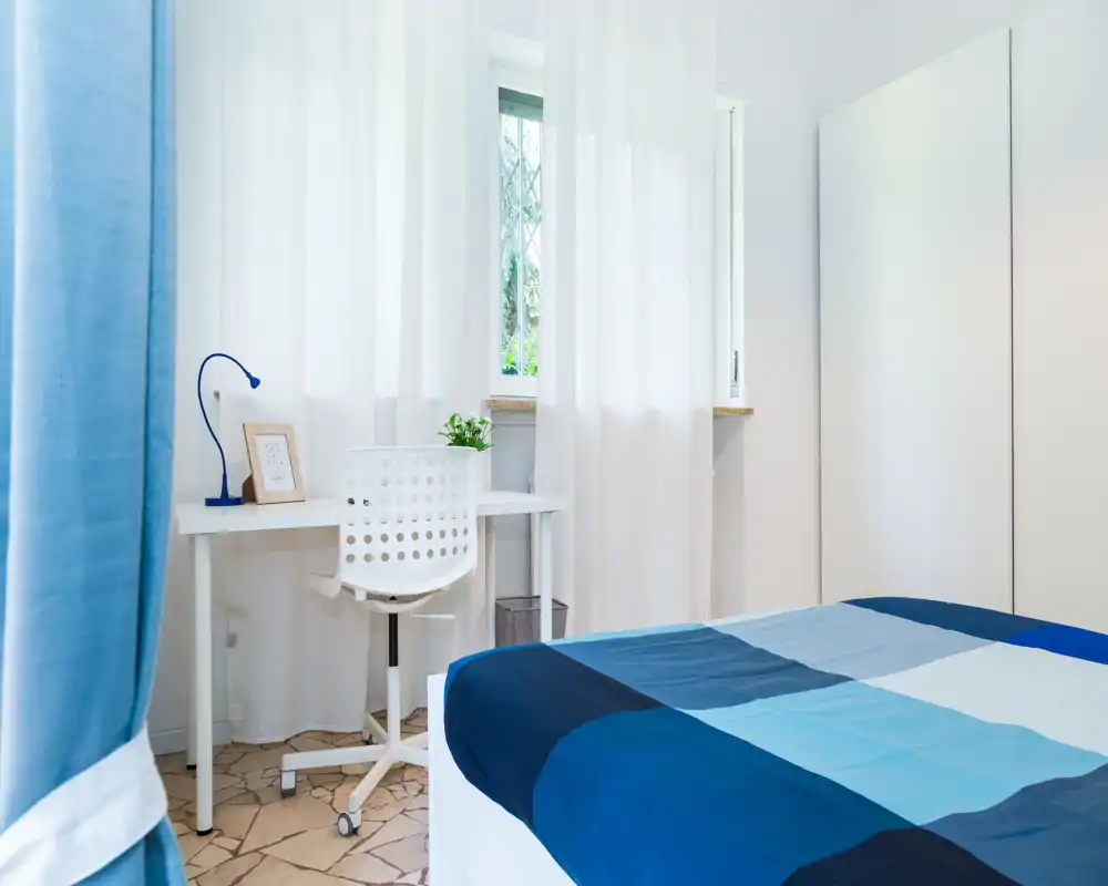 Camera da letto luminosa e ordinata con copriletto blu a quadri, una piccola scrivania e sedia vicino alla finestra e un armadio minimal. La luce naturale e l’arredamento coordinato creano un'atmosfera calma e accogliente ideale per affitti brevi.